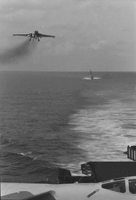 "7th Fleet" A view of the open ocean. A low-flying airplane approaches the aircaft carrier from which the photo was taken. In the background a second aircraft carrier can be seen.