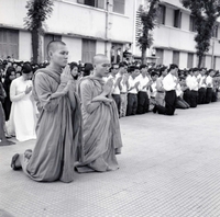 Two Buddhist monks kneeling and praying. A large group of Vietnamese people are kneeling and praying behind them.