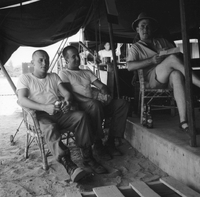 American men are sitting in chairs in the sand under a tent.