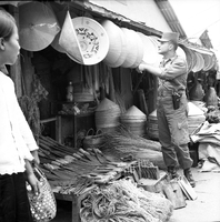 "Ca Mau" American soldier looking at Vietnamese hats hanging from market vendor with other goods at his feet.