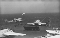 American men working on an aircraft carrier in open water. Four Naval planes are still on the runway, one with detached wings. One other have just taken off.