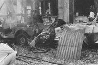 "Embassy Bombing" Two cars have collided in the steaming rubble. Two men enter a ruined building. A man crouches in the debri-ridden street to take a photograph.