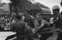 "Embassy Bombing" A crowd of people in the street. A military truck is parked in the middle of the street. Three men carry a body on a stretcher, looking to the left. A fourth man stands with one arm protectively over the body.