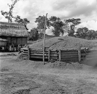 "Montagnards - Showplace Strat. Ham - 8-21-62 - In MTs - Faces - Blockhouses" A dirt mound contained by a wooden fence, near some huts.