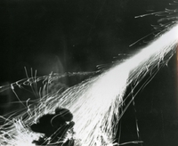 "And the Rockets Red Glare. ln the seemingly endless defense of the freedom of man, rockets continue to play a key role. As independence day 1967 approaches, the seventh fleet rocket firing ships the USS Clarion River contributes to the fireworks display seen constantly by the American fighting men serving in Vietnam. The Clarion River has been supporting allied forces ashore in the Republic of Vietnam." A long exposure photograph of sparks in the sky.  There is a silhouette in the foreground, which may be a man.