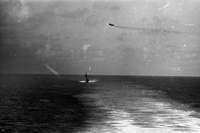 "7th Fleet" A view of the open ocean. An aircraft carrier and an ascending plane can be seen in the background. A wide strip of water in the foreground is rough in the wake of a second ship, out of view.
