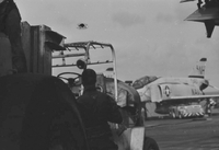 "7th Fleet" An American man running a large piece of machinery. A second man can be seen climbing the machine in the upper left. A Naval plane can be seen in the background. There is a small hole in the photograph above the machine.