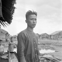 "VN Life", a potrait of a Vietnamese man standing on a dirt village road. There are two children in the background.