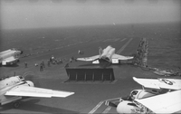 American men working on an aircraft carrier in open water. Four Naval planes are still on the runway, one with detached wings.