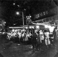"Sarawak." Night market in a Sarawak street. The market is crowded with people, especially women in fashionable dresses. The signs in the market are largley in Chinese characters. From scrapbook page 13.