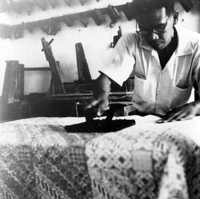 "Indonesian Arts and Crafts Negs [Negatives]." An Indonesian man stamps wax onto fabric to make batik. There is wooden furniture stacked against a wall in the background. From a scrapbook page 10.