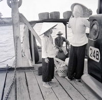 A Vietnamese woman and child stand on a dock with some baskets. A man is sitting in a boat docked there.