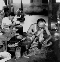 "Indonesian Arts and Crafts Negs [Negatives]." Indonesian men sit on the floor, spread apart in a large room filled with hammers, anvils, vices, and other metalworking tools. The man sitting closest to the camera seems to be shaping a spoon or cooking tool. From a scrapbook page 10.