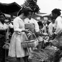 "Korea-Negs 181-3(1)-182-3(1) 4.Seoul Mkt [Market]" Two Korean women and a child stand at a market stall buying green onions. One woman is wearing western dress and the other woman is in traditional clothes. Most of the vendors in the photo appear to be older women. From scrapbook page 23.