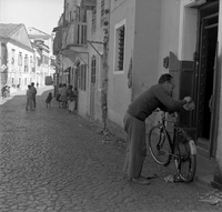 "Macau" A man leans on his bike talking with someone through a doorway, as a group of children are seen in the background further down the road.