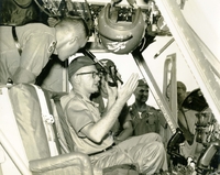 "Loi, Vietnam - Secretary of the Army Stanley R. Resor (seated) is briefed on the operation of the gun sight of a Huey helicopter by officers of the 11th Aviation Battalion. The commanding officer of the 11th, Lt. Col. Lauterback (glasses on lapel) stands at right. Other officers are unidentified. Secretary Resor was visiting combat and support units in Vietnam." An American pilot sits in a cockpit and talks to a group of four other airmen.