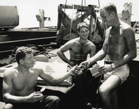 "Lunch Break - Members of Hatch Team #9, Seamen Charles C. Shearon and Robert J. Kinney, Jr., and Boatswain's Mate 2C Robert J. Ely, take mid-day, 30-minute lunch break topside on merchant vessel they are offloading. The break helps to split-up 12-hour work shift. Combat 'C-rations' is the menu for lunch." Three American Navy sailors sit together, either on a dock or on the deck of a ship. There is a box labelled "meal combat individual" behind them, and the sailors appear to be dividing up the rations inside the box. One holds an opened can and another holds a small box.