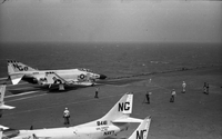 Men working on an aircraft carrier in open water. Three Naval planes sit on the runway. A man slightly right of center is also taking a photograph.