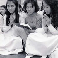 Three Vietnamese women sit cross legged and pray. The woman in the middle is holding a book.