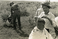 "Pleiku, Vietnam (Army IO)- PFC David J. Walker, a member of the Army's 173rd Airborne Brigade, from Montreal, Canada gets his "white sidewall" Airborne haircut while the trooper in the background pays the pretty cashier for his haircut." A Vietnamese man cuts an American soldier's hair. In the background, an American soldier hands a Vietnamese woman something while she sits on the ground.