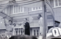 Nguyá»…n KhÃ¡nh holds something above his head as Robert McNamara stands in the background. They are standing on a stage with microphones in front of them. Henry Cabot Lodge, Jr., appears to be standing off to the side.