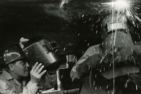"Running A Bead - Mr. Melvin Werner (left), a welder with the U.S. Naval Shipyard, Peral Harbor, Hawaii, keeps a close eye on a Vietnamese welder running a bead with an arc-welder on the underside of a medium landing craft (LCM) at the Saigon Naval Shipyard. Mr. Werner and three other welders from the Naval Shipyard in Pearl Harbor, currently on a six weeks tour under the United States Military Assistance Program, are teaching welding techniques to Vietnamese welders at the Saigon Naval Shipyard. They will return to Pearl Harbor on 12 December." An American man watches a Vietnamese man welding.