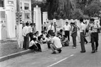 "Unidentified", a group of Vietnamese men gather and sit on the curb outside a gated building. They appear to be part of a protest as they have a big sign with them.
