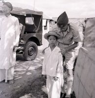 An American soldier talking to a young Vietnamese child. There is a Jeep and a Vietnamese man in the background.