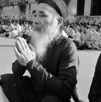 Original title: "VN Life", The photo is of a Buddhist religious service, which may have been part of the anti-Diem Buddhist Crisis protests. An old Vietnamese man in traditional clothes sits on the ground in the foreground praying. There is a large crowd of young people praying behind him, and Buddhist monks stand along a wall in the background.