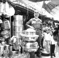"Ca Mau" American soldier standing behind some metal objects with curious children standing beside him for the photo.