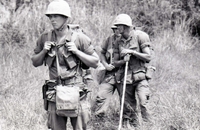 Three American soldiers walking through a field in a line. The soldier in the middle is holding a walking stick.