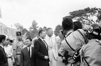 Robert McNamara and Henry Cabot Lodge Jr walk in front of a crowd holding banners and flags. There is press in front of him taking photos.