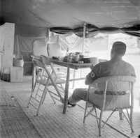 "QN + Train ". Picture of a soldier in sitting on a chair, with a table filled with food ? In front of time, ina atent