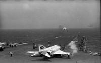 Men working on an aircraft carrier in open water. Several Naval planes are still on the runway. One has just taken off.