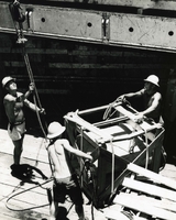 "The Heat is On - George Nolan, Boatswain's Mate 1C, assists Robert J. Ely, Boatswain's Mate 2C, and Bobby R. Loftis, seaman, members of his Hatch Team #9, in slinging cargo in the 130 degree heat of #1 hold aboard S.S. Sir John Franklin. The men alternate between topside and below decks every 20-30 minutes due to extreme heat." American sailors tie up a crate of cargo with rope, preparing it for loading into a ship. They appear to be on a wooden dock.
